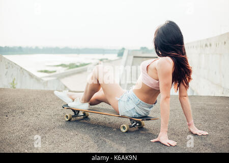 Schöne junge Mädchen mit Tätowierungen sitzt auf Longboard bei schönem Wetter Stockfoto