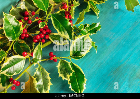 Ein Weihnachten Hintergrund mit Stechpalme Blätter und Beeren auf ein leuchtend blaue Textur mit Girlande Lichter und Kopie Raum Stockfoto