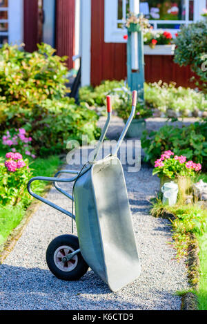 Metall Schubkarre auf Kies Lane seitig von Blumenbeeten. Infrarotkabine im Hintergrund. Stockfoto