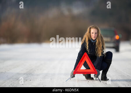 Junge Frau ein Warndreieck Einrichtung und Unterstützung gefordert, nachdem ihr Auto in der Mitte des nirgendwo an einem eiskalten Wintertag ist kaputt Stockfoto