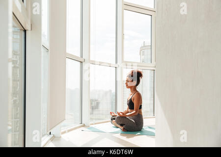 Junge afrikanische Fitness lady in Sportkleidung sitzen im Lotussitz und suchen Kamera zu Hause Stockfoto