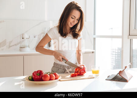 Attraktive casual Frau schneiden Paprika auf einem Holzbrett für einen Salat aus einer Rezeptur auf Tablet Computer in einer Küche Stockfoto