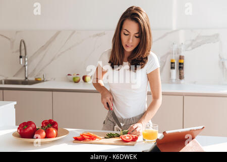 Acht junge Frau Schneiden von Gemüse auf einem Holzbrett und Tablet Computer beim Kochen in der Küche Stockfoto