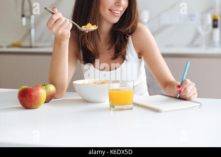 Zugeschnittenes Bild eines lächelnden Frau isst Müsli mit Orangensaft und Notizen in Notepad am Küchentisch Stockfoto