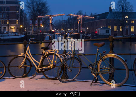 Abend geschossen von einem malerischen Brücke über einen Kanal in Amsterdam im Winter mit Schnee und schöne Lichter, mit einigen Fahrräder im Vordergrund. Stockfoto