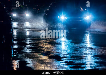 Nacht der Verkehr in der Stadt. Autos fahren durch große Wasserpfützen mit blauen Scheinwerfer. Stockfoto