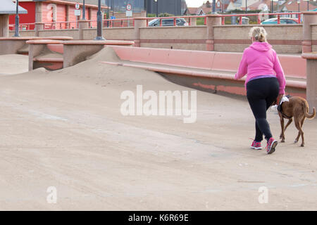 Einsame weibliche Wandern einen Hund Rhyl Promenade im Norden von Wales mit dem Driften sands Aufbau gegen die Promenade als Ergebnis Sturm Aileen, Denbighshire, Wales, Großbritannien Stockfoto