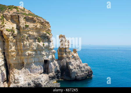 Ein Kalkstein Klippe mit einer kleinen Höhle und zwei short stacks, entlang der maltesischen Küste gefunden. Das Meer ist ruhig mit einem Handelsschiff Ferne. Stockfoto