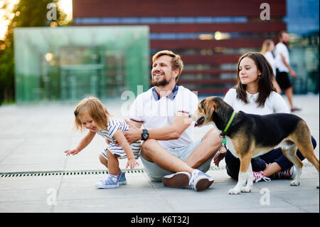 Nette junge Familie ruht sich auf dem Boden sitzend. Mann, Frau, ihrer kleinen Tochter und einen Hund. Kind bricht spielend aus Daddy's Händen. Vater che Stockfoto