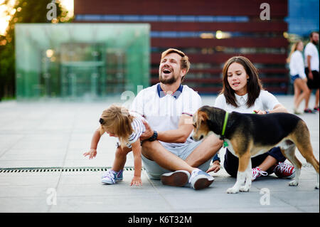 Nette junge Familie ruht sich auf dem Boden sitzend. Mann, Frau, ihrer kleinen Tochter und einen Hund. Kind bricht spielend aus Daddy's Händen. Vater che Stockfoto