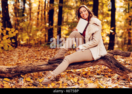 Schöne lächelnde Mädchen im Wald im Herbst. Sie sitzt auf dem Baum, Buch und denken. Stockfoto