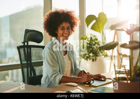 Portrait von schönen weiblichen Designer sitzen an Ihrem Schreibtisch bei der Kamera schauen und lächeln. Afro-amerikanische junge Frau von ihrer kreativen Arbeitsplatz. Stockfoto