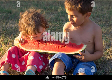 Kleine Mädchen und Jungen essen riesige Scheibe Reifen Sommer Wassermelone, sitzen auf der Wiese bei Sonnenuntergang. Stockfoto