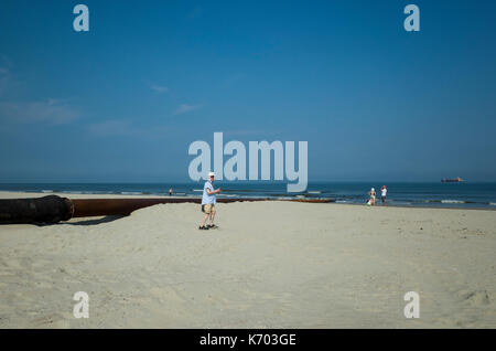 Am Strand, Langeoog. Deutschland. Deutschland. Touristen sind noch in der Lage zu Fuß am Strand entlang, während der aktiven Strand Umbau, Strandaufschüttung genießen. Zwei der Handelsschiffe, die den Sand Pumpen werden entlang der Rohre können am Horizont gesehen werden. Es ist ein Sonniger Tag mit kaum sichtbaren Cloud gegen den blauen Himmel. Stockfoto