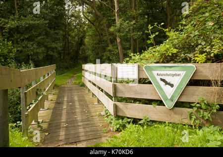 Am Wald, Langeoog. Deutschland. Deutschland. Ein Fußweg und kleine Holzbrücke in die natürlichen Wälder führt. Das Zeichen, 'Naturschutzgebeit', für die natürliche befindet sich auf der hölzernen Zaun führenden auf zur Brücke über einen Graben und in das Holz positioniert. Dappled Sonnenlicht erzeugt Muster auf den Boden. Stockfoto