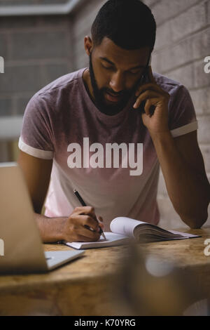 Junger Mann Gespräch am Handy beim Schreiben auf Tagebuch im Cafe Stockfoto