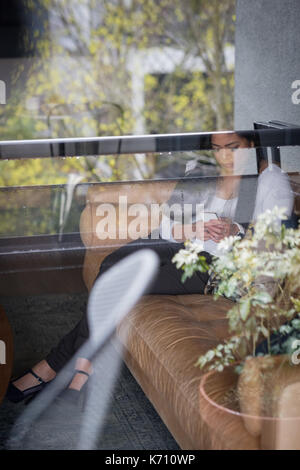 Junge Geschäftsfrau mit Telefon im Büro durch Glas gesehen Stockfoto