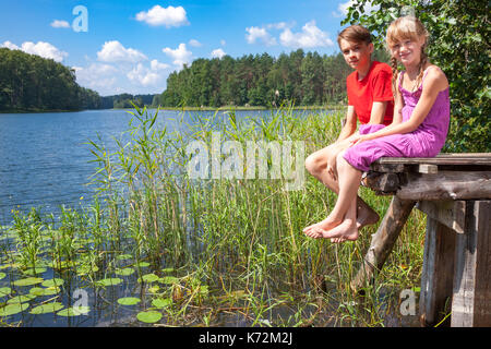 Teenager Boy und elementare Alter Mädchen sitzen auf einem hölzernen Pier von einem Wald See genießen Sommer Tag im Freien. Stockfoto