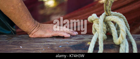 Fisherman's Hand mit Fischernetz im Hintergrund. Nass- und faltige Hand stützte sich auf ein hölzernes Boot Zaun. Fisherman's Hand neben Seil gebunden. Stockfoto