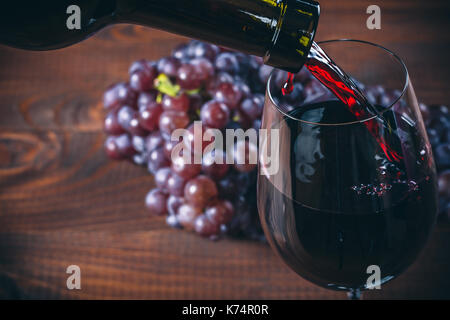 Gießen Rotwein ins Glas mit einem Strauß roter Trauben gegen Holz- Hintergrund Stockfoto