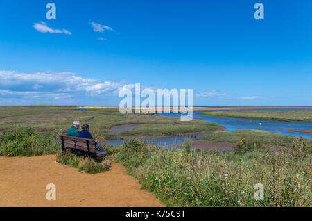 Senior Paar sitzt auf einem Strand mit Blick auf die Küste bei Thornham Hafen, North Norfolk, Großbritannien Stockfoto
