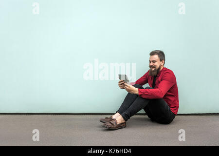 Arbeiten im Freien, sitzt auf dem Boden. Junge Erwachsene bärtigen Geschäftsmann arbeiten im Tablet- und lächelnd. Studio gedreht. Auf grauem Hintergrund. Stockfoto