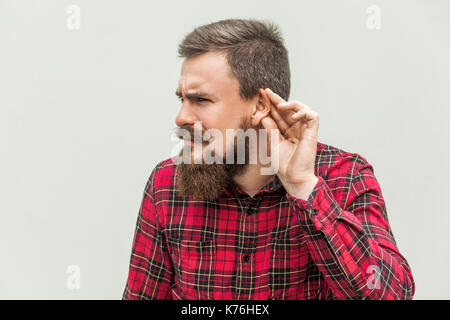 Ich kann Sie nicht hören. Junge Erwachsene bärtiger Mann hören. Studio shot, grauer Hintergrund Stockfoto