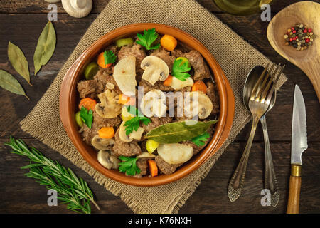 Foto von einem fleischragout in einem irdenen Schüssel, der mit Zweigen von frischem Rosmarin, Lorbeer und Pilze, und eine hölzerne Schöpfkelle mit Pfefferkörnern, shot f Stockfoto