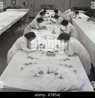 Eine historische Gruppe von Frauen aus den 1950er-Jahren, die auf langen Tischen mit Pinsel Entwürfe auf gewebte Leinentücher gemalt haben, Nordirland. Irisches Leinen ist ein Naturstoff mit langer Tradition, der aus der Leinpflanze (Unkraut) hergestellt wird, die natürlich auf argem Land und bei Regen wächst. Stockfoto