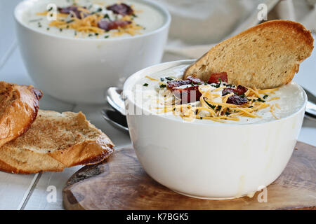 Heiße Kartoffelsuppe mit Sauerrahm, Cheddar Käse und Schnittlauch mit in Scheiben geschnittenen, geröstetes Brot. Extrem flache Tiefenschärfe mit selektiven Fokus auf Schüssel in Stockfoto