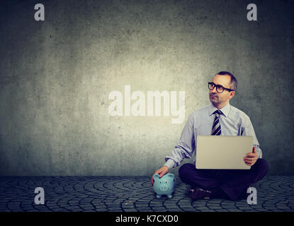Nachdenklich business Mann mit Laptop und piggy Bank sitzen auf einem Stock Stockfoto