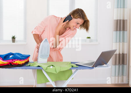 Junge Frau mit elektrischen Bügeleisen Gespräch am Handy und Laptop Suchen Stockfoto