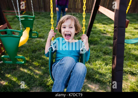 Portrait von glücklichen Mädchen schwingen auf Spielplatz mit Mutter im Hintergrund Stockfoto