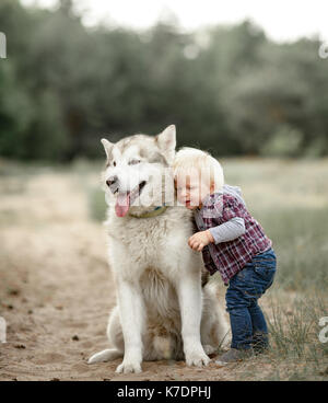 Little Boy steht, Umarmungen sitzen Malamute Hund und Knuddel ihn für Spaziergang entlang der Forststraße. Stockfoto