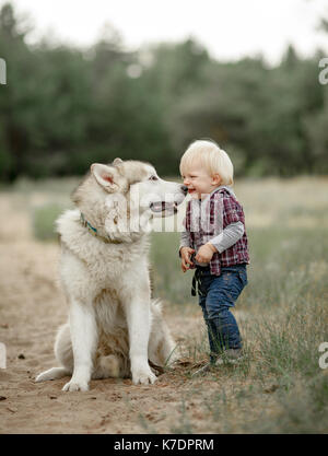 Kleiner Junge steht in der Nähe sitzen malamute Hund auf dem Spaziergang entlang der Forststraße und lächelt. Stockfoto