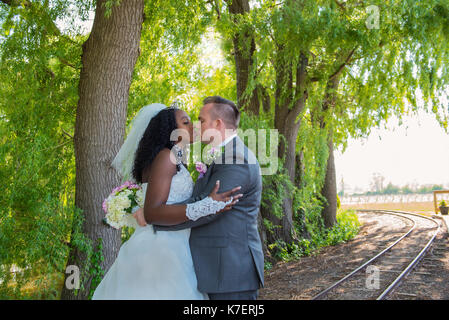 Brautpaar küssen auf eine Bahn unter Bäumen, interracial Hochzeit Stockfoto