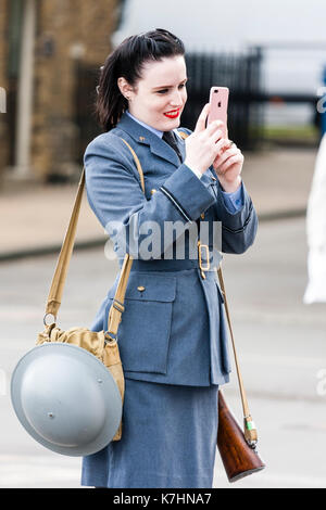 England, Chatham Dockyard. Veranstaltung, Gruß zu den 40er Jahren. Frauen in der RAF Uniform mit Gewehr slang über die Schulter und Zinn Helm hängen von anderen Schulter. Ständigen, lächelnd und mit dem iPhone zu. Stockfoto