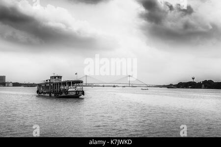 Öffentliche Fähre überquert den Hooghly River an einem bewölkten Monsunmorgen mit Blick auf die neue Howrah Brücke im Hintergrund in Kalkutta, Indien. Stockfoto