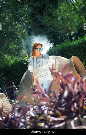 Junge Frau vaping mit Sonnenbrille in einer natürlichen Szene. Stockfoto