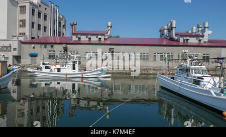 Horizontale Schuß des weißen Fischerboot Position heraus zum Meer von Otaru Kanal mit Factory im Hintergrund. Reflexionen von Gebäuden und Boote in blue water Stockfoto