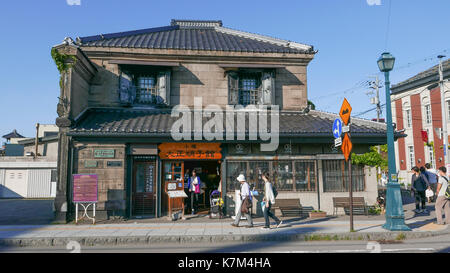 Käufer vorbei gehen traditionelle Storefront in Otaru Japan. Historische Gebäude aus Stein mit touristischen Informationen Zeichen vor. Orange Holz- Kanji Stockfoto