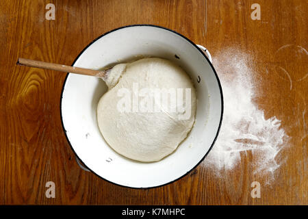 Steigende Hefeteig mit Holzlöffel in Weiß Emaille metall Schüssel auf hölzernen Tisch von oben. Verschüttetes Mehl. Stockfoto