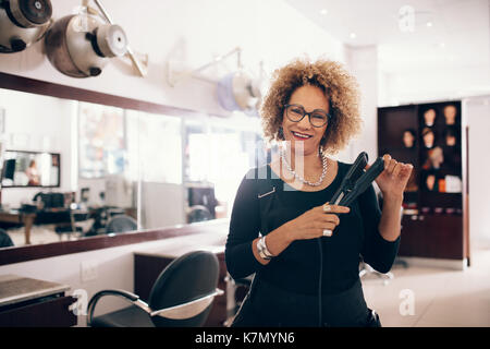 Lächelnd friseur Holding eine haarglättung Werkzeug. Gut ausgestattete Beauty Salon mit professionellen Friseur. Stockfoto