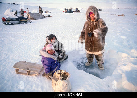 Inuit Mädchen im Alter von 4 und 11, zusammen mit dem Besucher, durch die Bohrung. Alle außer das jüngere Mädchen, einschließlich anderer Besucher und Inuit ältesten im Hintergrund, sind im traditionellen Caribou haut Kleidung. Außerhalb Baker Lake, Nunavut, Kanada. Stockfoto