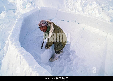 Inuit ältester Mann, Mitte der 60er Jahre, in der modernen Winter Kleidung, Schnitte Eisblöcke mit Messer und schneidet sie während Gebäude im traditionellen Iglu. Stockfoto