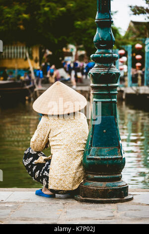 Native Frau mit Strohhut sitzt am Ufer des Thu Bon Fluss, Hoi An, Vietnam Stockfoto