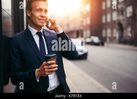 Junge executive über sein Handy und trinken einen Kaffee, während gegen eine Wand auf einer Straße der Stadt am Morgen schiefen Stockfoto