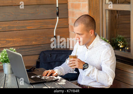 Hübscher junger Geschäftsmann Arbeiten am Notebook während der Sitzung einen Holztisch in modernen Kaffee Shop Interior, junger Mann trinken Kaffee im Cafe und Usi Stockfoto