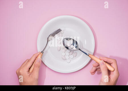 Draufsicht auf die Hände des Mannes am Esstisch mit Messer und Gabel vor Gericht mit Pillen über rosa Hintergrund. Stockfoto