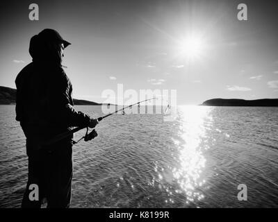 Fischer prüfen Angeln und Köder auf der Stange, selbst vorbereiten und weit in das ruhige Wasser locken werfen. Fischer Silhouette bei Sonnenuntergang Stockfoto